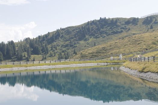 Scenic view to small alpine lake and big mountains with glacier in sunlight. Awesome green landscape with blue mountain lake among mosses in green highland valley in sunny day. Wonderful scenery.