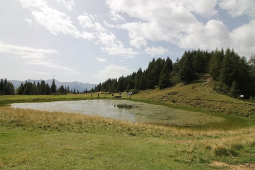 Scenic view to small alpine lake and big mountains with glacier in sunlight. Awesome green landscape with blue mountain lake among mosses in green highland valley in sunny day. Wonderful scenery.