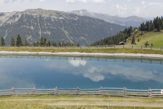 Scenic view to small alpine lake and big mountains with glacier in sunlight. Awesome green landscape with blue mountain lake among mosses in green highland valley in sunny day. Wonderful scenery.