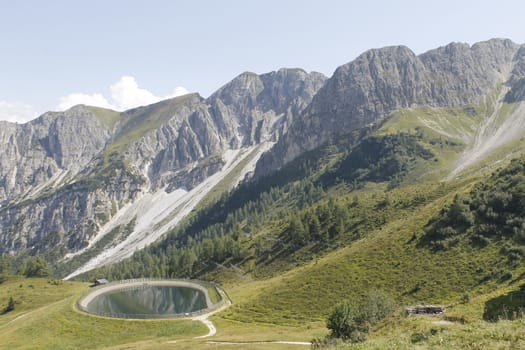 Scenic view to small alpine lake and big mountains with glacier in sunlight. Awesome green landscape with blue mountain lake among mosses in green highland valley in sunny day. Wonderful scenery.