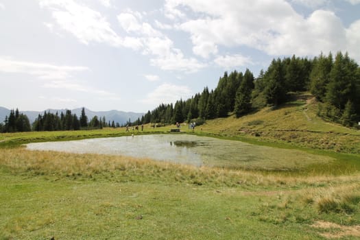 Scenic view to small alpine lake and big mountains with glacier in sunlight. Awesome green landscape with blue mountain lake among mosses in green highland valley in sunny day. Wonderful scenery.