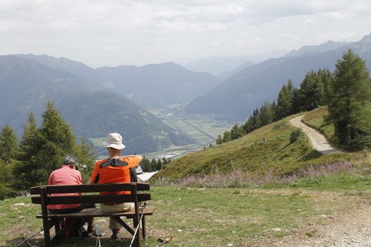 A  couple is sitting on a bench and looking at the mountains.