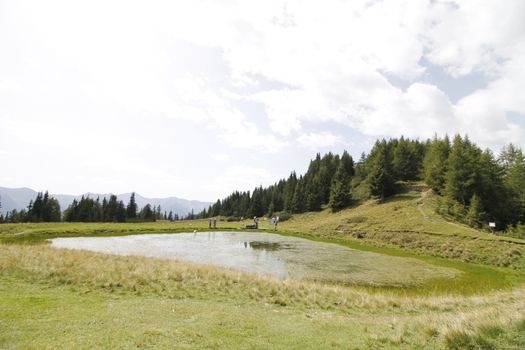 Scenic view to small alpine lake and big mountains with glacier in sunlight. Awesome green landscape with blue mountain lake among mosses in green highland valley in sunny day. Wonderful scenery.