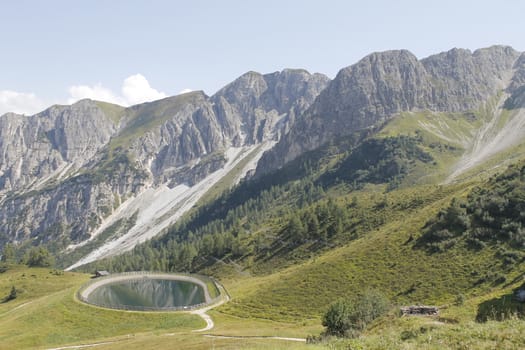 Scenic view to small alpine lake and big mountains with glacier in sunlight. Awesome green landscape with blue mountain lake among mosses in green highland valley in sunny day. Wonderful scenery.