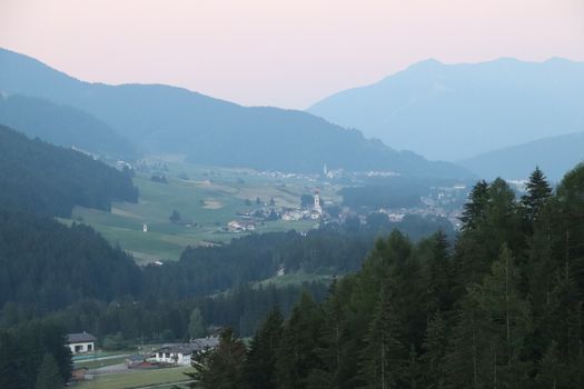 Aerial view of huge valley of the mountains of Italy, Trentino, green meadows, Slopes with green spruce trees, Dolomites on background, The town in the bottom of a valley