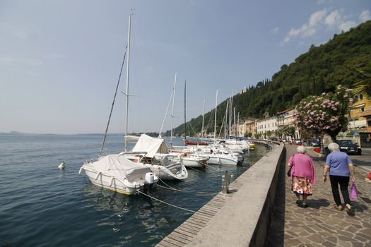 View on sailing yacht and boats parked on crystal clear blue water of amazing lake Garda and Toscolano Maderno cityscape on the background