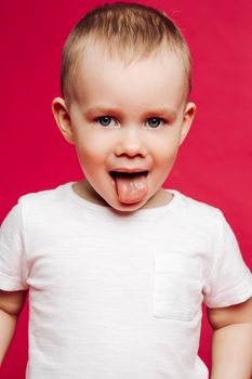 Portrait of little boy with blue eyes, showing tongue out, playing at studio. Kid in white t-shirt stick out and making face, posing, looking at camera. Pink studio background. Concept kids fashion.