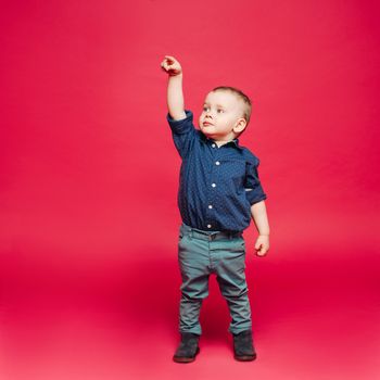 Studio portrait of little stylish boy with blue eyes gesturing and pointing up. Child wearing in shirt, playing at pink studio. Kids fashion.