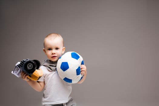 Studio portrait of little sweet boy posing at camera with different equipment of work, photo camera, foot ball. stylish kid in scarf and white t shirt at gray studio. Concept of choice profession.