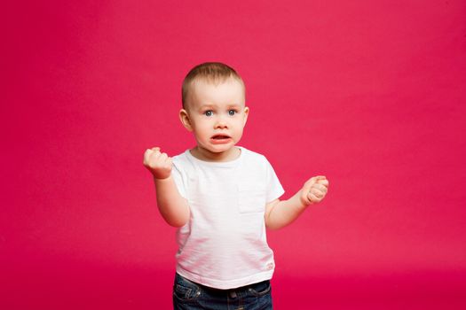 Emotionaly portrait of little boy shocked and surprised looking, gesturing by hands. Sweet fashionable child in white t shirt and jeans, playing and screaming at pink studio.