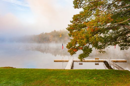 Scenic river edge and T-shaped pier under oak tree early morning fog just above the water level of Connecticut River with colorful fall foliage along both sides converging into distance.