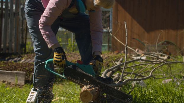 Mature woman cutting tree with an electric chain saw on the courtyard of a country house. Active and healthy lifestyle concept