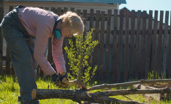 Mature woman chopping branches of a tree with ax in the yard of a country house. Active and healthy lifestyle concept