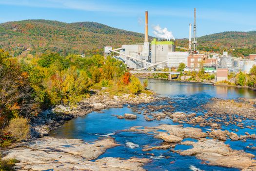 Idyllic environment stream and Rumford Falls conflicts with industrial factory surrounded by beautiful forested hills and autumn foliage colors.