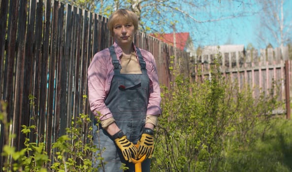 Portrait of mature blond caucasian woman with a shovel in her hands in the countryside garden. Sunny day, spring. Healthy and active lifestyle concept.
