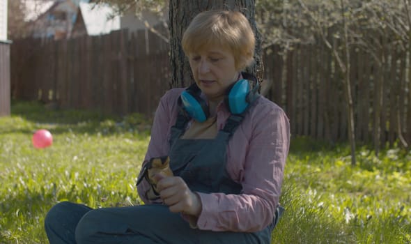 Mature woman sitting near the pine tree in the garden after work. She taking off protective gloves. Middle-aged caucasian woman in overalls in the yard at spring day