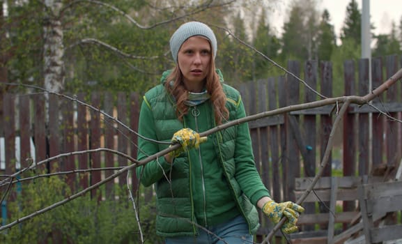 Young beautiful woman collecting firewood in the courtyard of a country house. Woman carrying a tree branch for a bonfire. People activity and healthy lifestyle concept