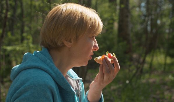 Close up portrait of mature blond woman eating sanwich in the forest. Snack during trekking. Healthy lifestyle concept