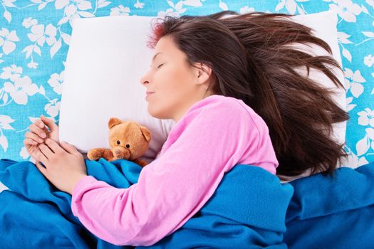 Close up of a beautiful young girl in pink pajamas sleeping in her bed, hugging a cute plush teddy bear. Sleep and relaxation concept.