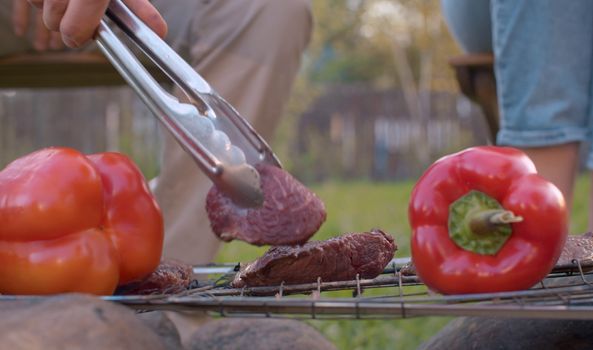 Close up hand putting piece of meat on the grill. Cooking of beef and vegetables on barbecue grill outdoor. Leisure, food, people and holidays concept. Top view