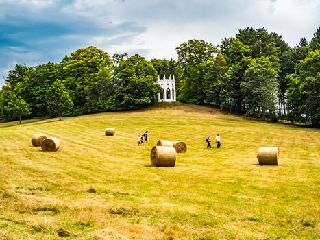 Painshill Park, Cobham Surrey, England. 4 August 2019. Painshill Park Surrey a family running across a yellow grass filed past hay bales with a white folly in the background.