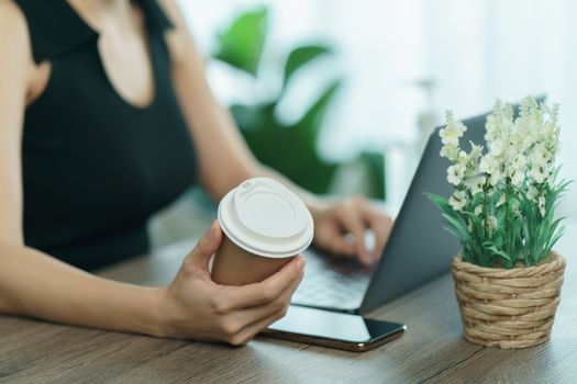 Young woman drinking coffee from disposable cup and using computer laptop on work desk.