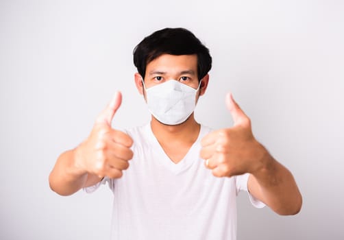 Asian handsome Man wearing surgical hygienic protective cloth face mask against coronavirus he show finger thumb for good, studio shot isolated white background, COVID-19 medical concept
