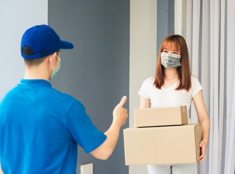 Asian delivery express courier young man giving parcel boxes to woman customer receiving both protective face mask and show thumbs up finger for good support sign, under curfew pandemic coronavirus