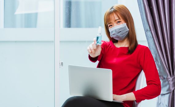 Work from home concept, Asian beautiful young woman smiling, student girl sitting on chair during working with laptop computer at home office and show alcohol spray sanitizing on hand