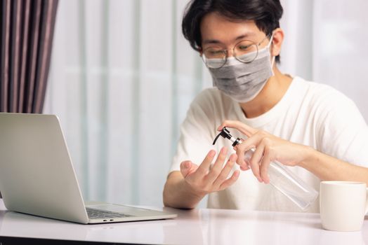 Asian business young handsome man glasses working from home office he quarantines disease coronavirus or COVID-19 wearing protective mask and cleaning hands with sanitizer gel on front laptop computer