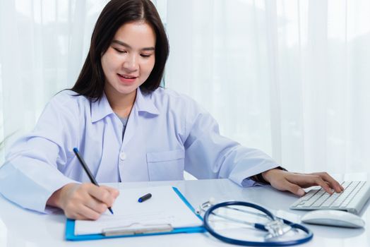 Asian doctor young beautiful woman smiling using working with PC desktop computer and her writing something on paperwork or clipboard white paper at hospital desk office, Healthcare medical concept