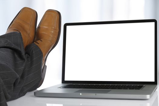 Businessman sits with his feet on the table next to a laptop with white screen.