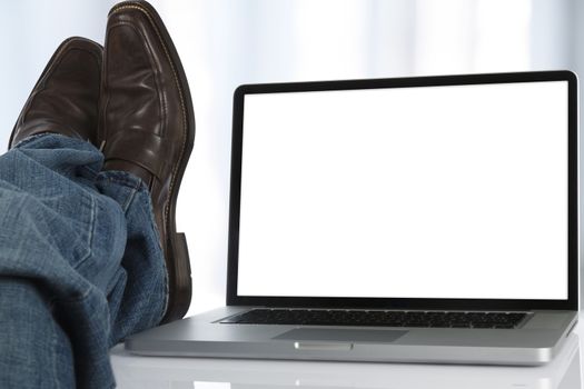 Man sits with his feet on the table next to a laptop with white screen.