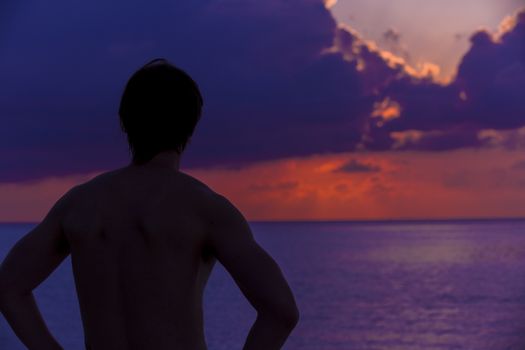 Beautiful idyllic seascape in The Maldives at sunset. A silhouette of the man is standing in front of the sun when it is getting darker and darker. The colors of the image are purple, blue and orange in a perfect contrast with the man in the beach.