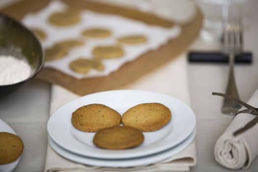 Plate with cookies in the table at front. In the back, out of focus, there are the natural ingredients to bake the cookies whick would be eggs, flour, water. The whole image is warm giving the message of cozyness.