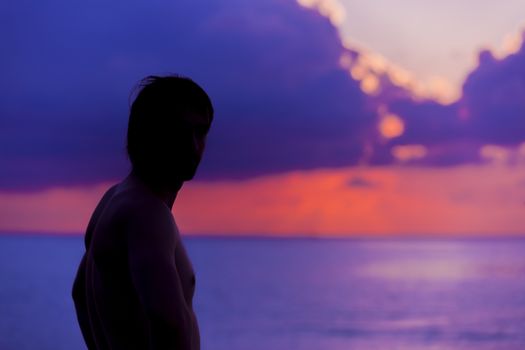 Beautiful idyllic seascape in The Maldives at sunset. A silhouette of the man is standing in front of the sun when it is getting darker and darker. The colors of the image are purple, blue and orange in a perfect contrast with the man in the beach.