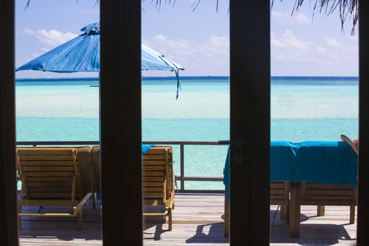 View from the inside of the bungalow through the window  in The Maldives. On the Deck there are deck chairs and beach umbrella. It is a beuatiful view of the sea, the water is clear, turquoise and in a strong contrast depending on the depth.