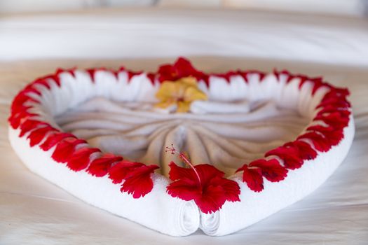 Towel forms a beautiful heart decorated with red flowers in a Luxury Resort room in Maldives. The heart is in the middle of the bed, the floor is wooden made.