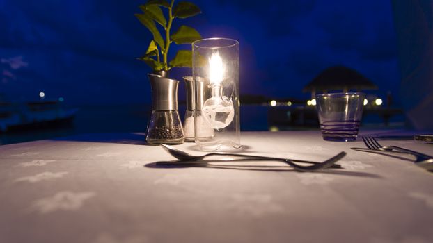 Table with candleand dinner setting in the beach. At background there are a boat and a bungalow over water. The set up here is a low key configuration.