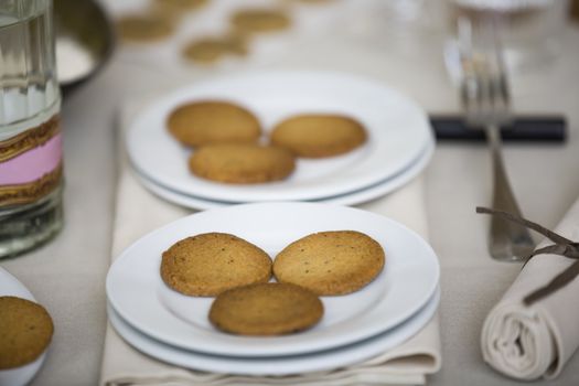Plate with cookies in the table at front. In the back, out of focus, there are the natural ingredients to bake the cookies whick would be eggs, flour, water. The whole image is warm giving the message of cozyness.