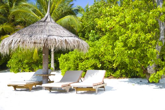 Three chairs and umbrella in the beach in Maldivess. The set is in the left of the frame, at background there is green vegetation and palm trees.