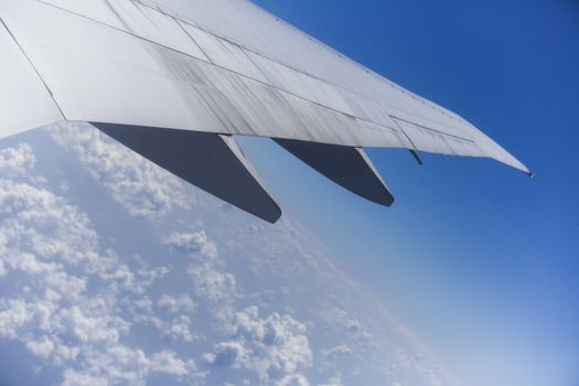 View from the right side of an aircraft of the wing, the plane is ready to land. It is a sunny day with a clear blue sky with clouds beneath it.