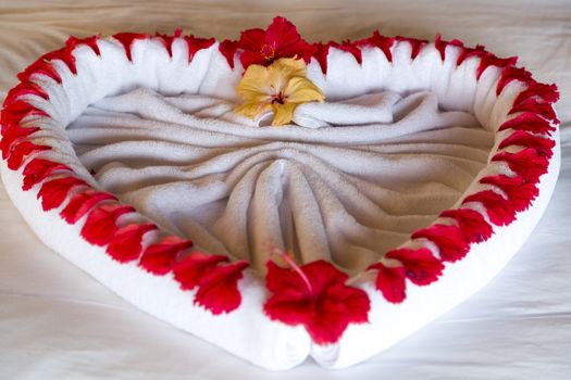 Towel forms a beautiful heart decorated with red flowers in a Luxury Resort room in Maldives. The heart is in the middle of the bed, the floor is wooden made.