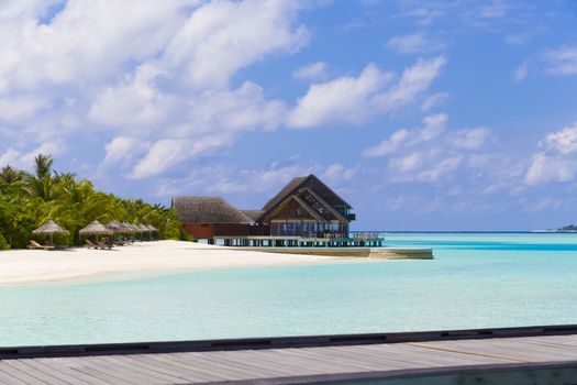 Beautiful View from the Beach with Jetty in Front and a Restaurant at Background. In the Beach there are beautiful green plants and deck chairs and beach umbrellas.