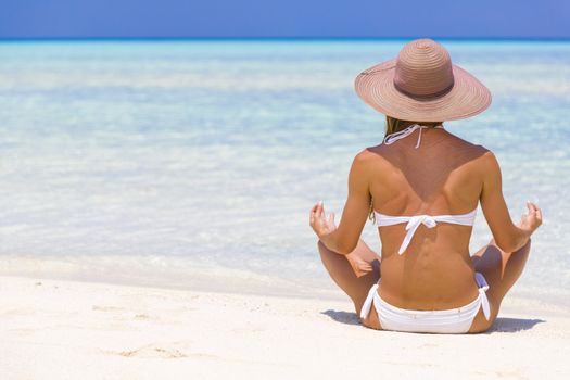 Young lady sitting on the shore in Maldives with straw hat meditating.