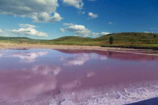 Pink salt basin in the front and green mountains in background with a clear blue sky. 