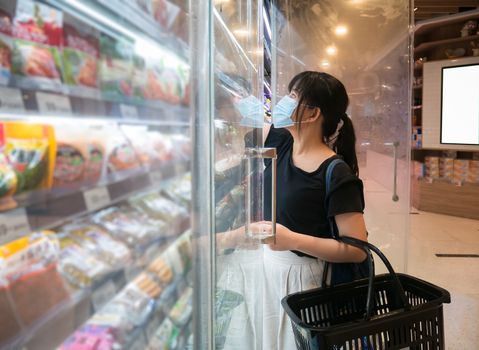 Asian women are shopping at the grocery store, holding baskets and wearing a health mask to prevent infection. She's opening the fridge to pick up the goods in lifestyle shopping concept