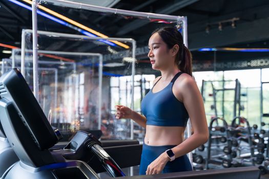 Women are running on a treadmill at the gym. Asian women wear fitness cloth, are exercising and have a barrier to prevent aerosols from exercising to keep social distancing