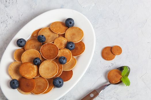 Trendy homemade breakfast, pancake cereal, mini pancakes on white plate on white concrete background with spoon and blueberry, leaves of lemon balm and peppermint