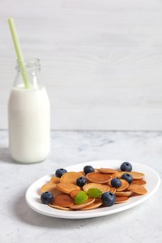 Trendy homemade breakfast, pancake cereal, mini pancakes on white plate on white concrete background with fork and blueberry, leaves of lemon balm and peppermint and bottle of milk